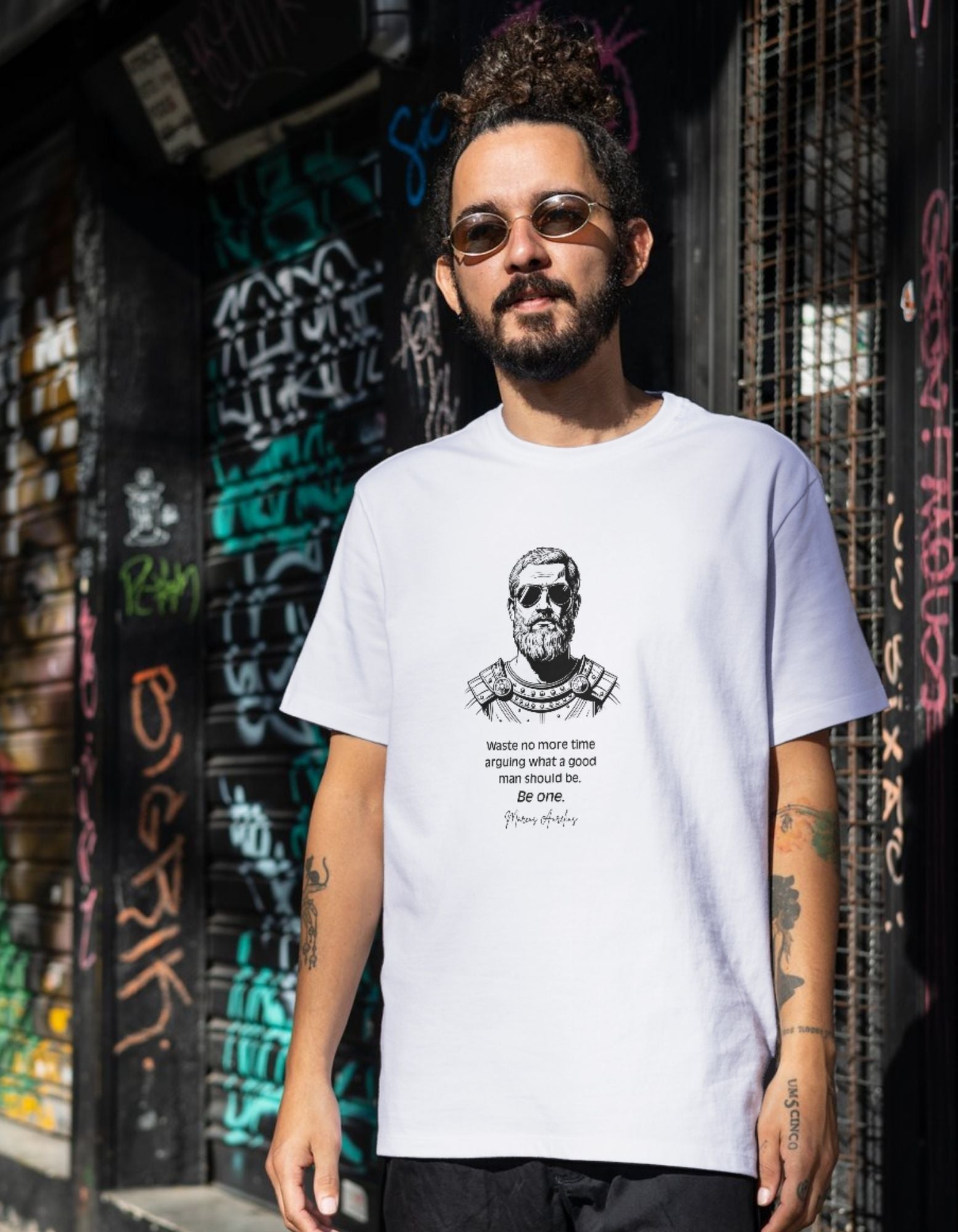 Man wearing a white t-shirt with a graphic design and text, standing in front of a graffiti-covered wall.